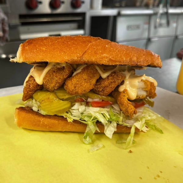 A fried shrimp po'boy sandwich from Noonie's Place, with lettuce, pickles, and sauce on a yellow paper.